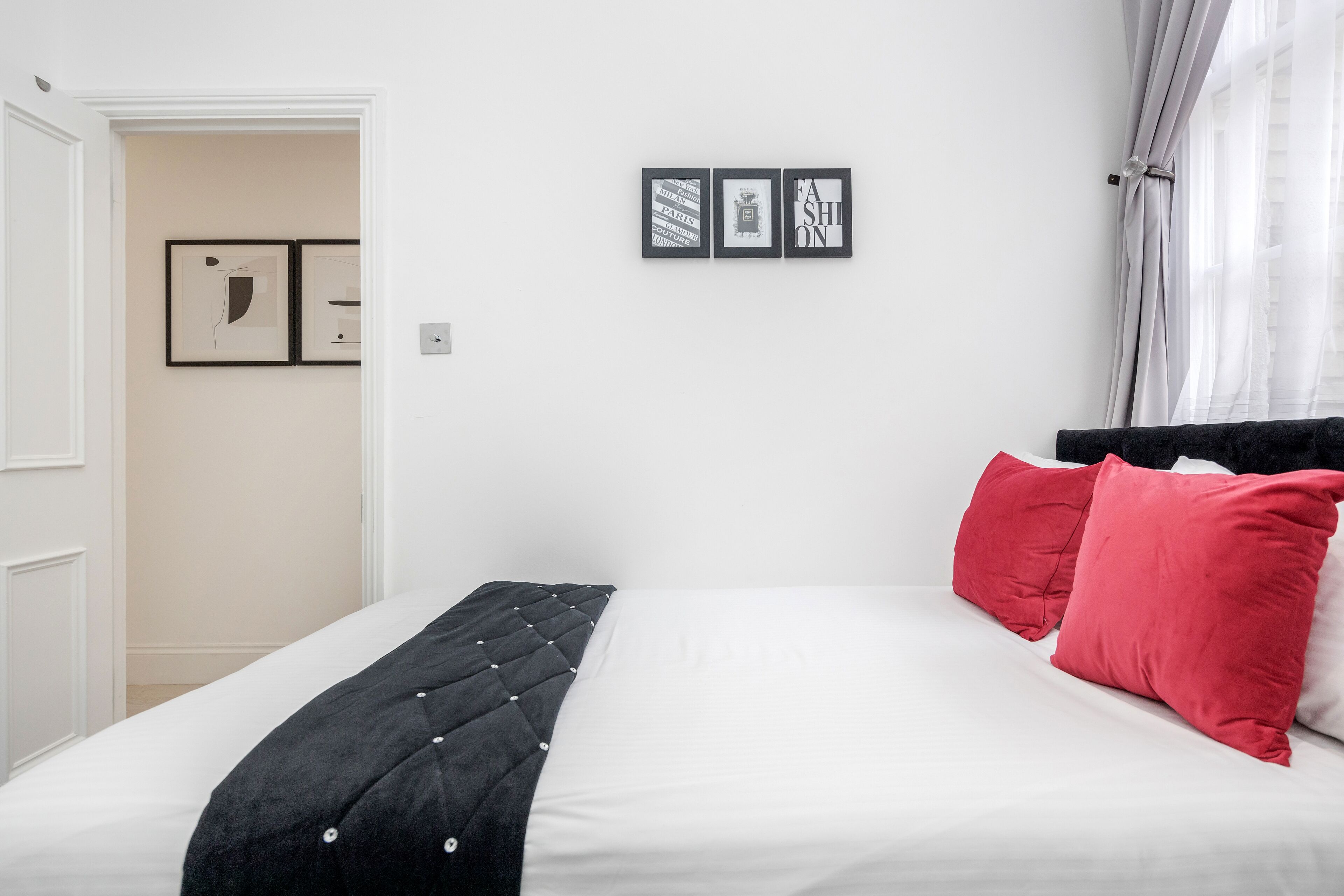 Een net opgemaakt bed met een zwart hoofdbord, witte beddengoed en rode kussens. Daarboven hangen drie ingekaderde foto’s aan de muur.