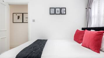 Een net opgemaakt bed met een zwart hoofdbord, witte beddengoed en rode kussens. Daarboven hangen drie ingekaderde foto’s aan de muur.