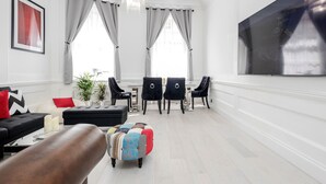 Uma sala de estar moderna com um sofá preto, almofadas vermelhas, um banquinho colorido e um espaço de jantar com cadeiras pretas e um lustre.