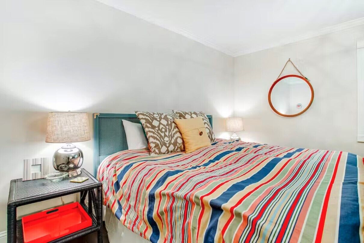 A bedroom with a striped bedspread, a round mirror on the wall, and a nightstand with a lamp and a red storage drawer.