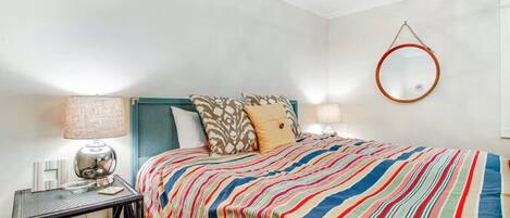 A bedroom with a striped bedspread, a round mirror on the wall, and a nightstand with a lamp and a red storage drawer.