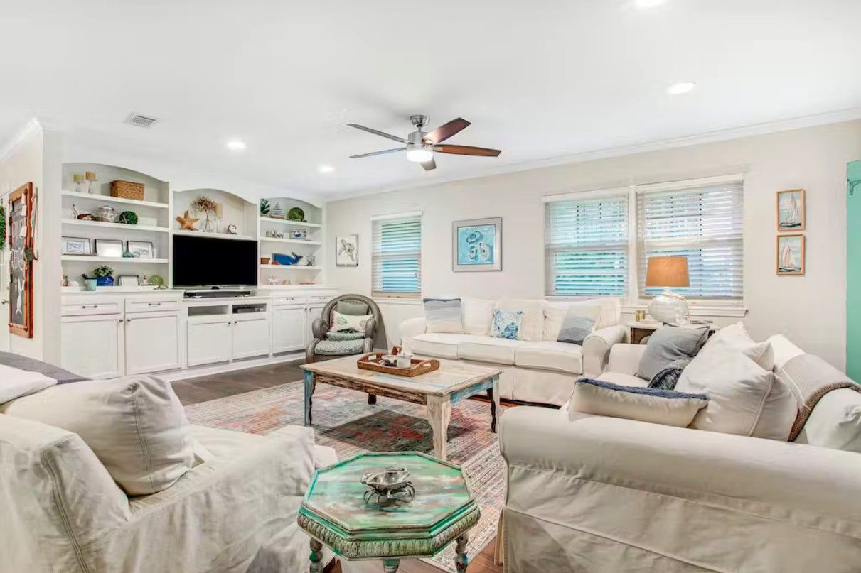 A modern living room with a white sofa, a coffee table, and a TV. There is a ceiling fan, a kitchen area with white cabinets, and a window with blinds.
