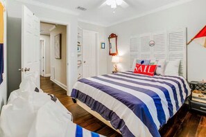 Une chambre avec une couvre-lit à rayures, un coussin orné d’une voile, une lampe de chevet et une fenêtre avec des volets.