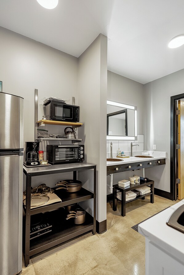 A compact kitchen with a microwave, toaster oven, and coffee maker on a shelf, a refrigerator, and a sink with a mirror above.