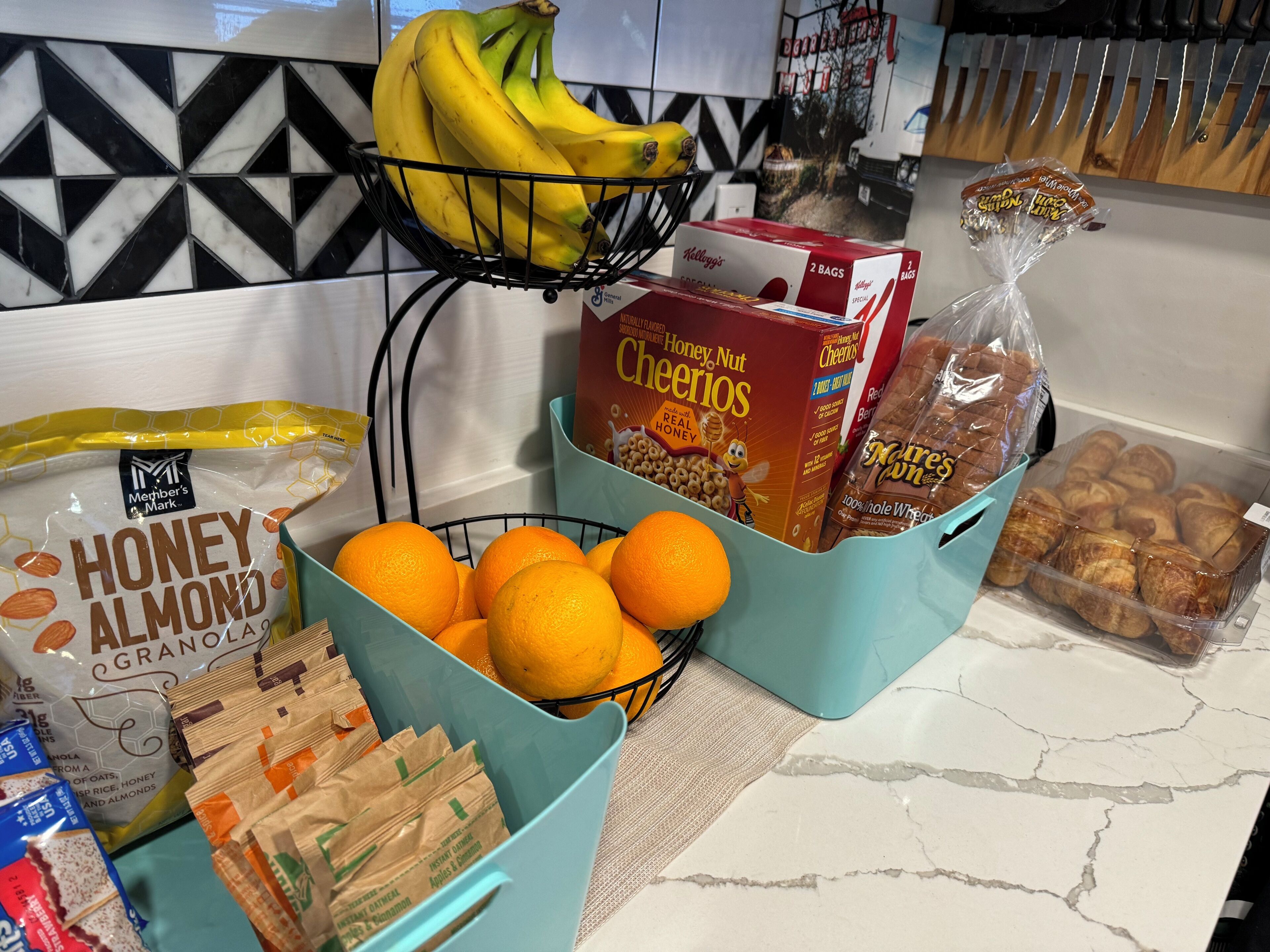 Eine Küchenarbeitsplatte mit verschiedenen Lebensmitteln, darunter Honig-Mandel-Granola, Orangen, Bananen, Cheerios und verpacktem Brot.