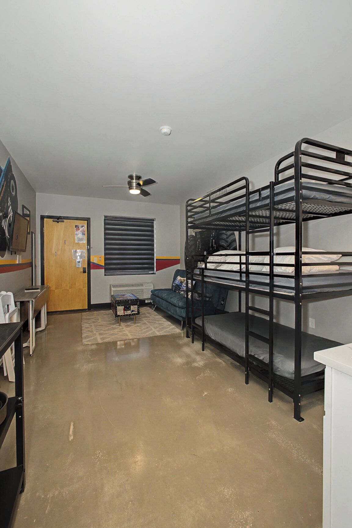A dormitory room with bunk beds, a ceiling fan, a window with blinds, a door, and a small table with chairs.