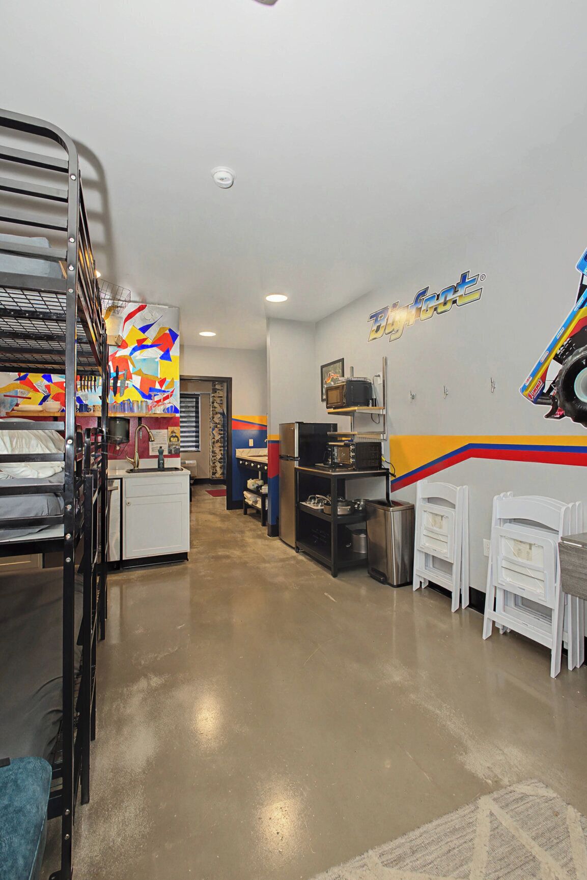 A room with bunk beds, a kitchenette, a dining area with white chairs, and a colorful abstract mural on the wall.