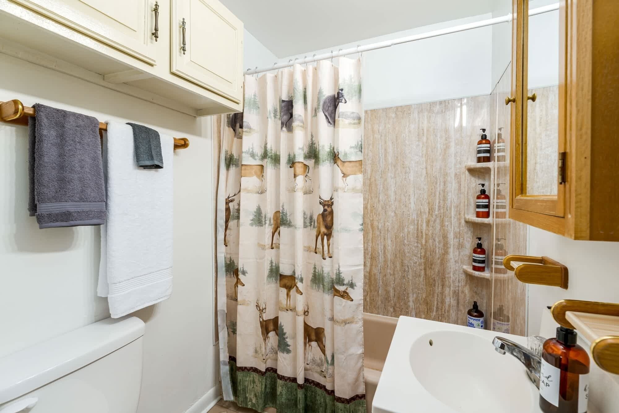 Un bagno con tenda da doccia raffigurante cervi, un lavandino bianco e articoli da toilette su uno scaffale.