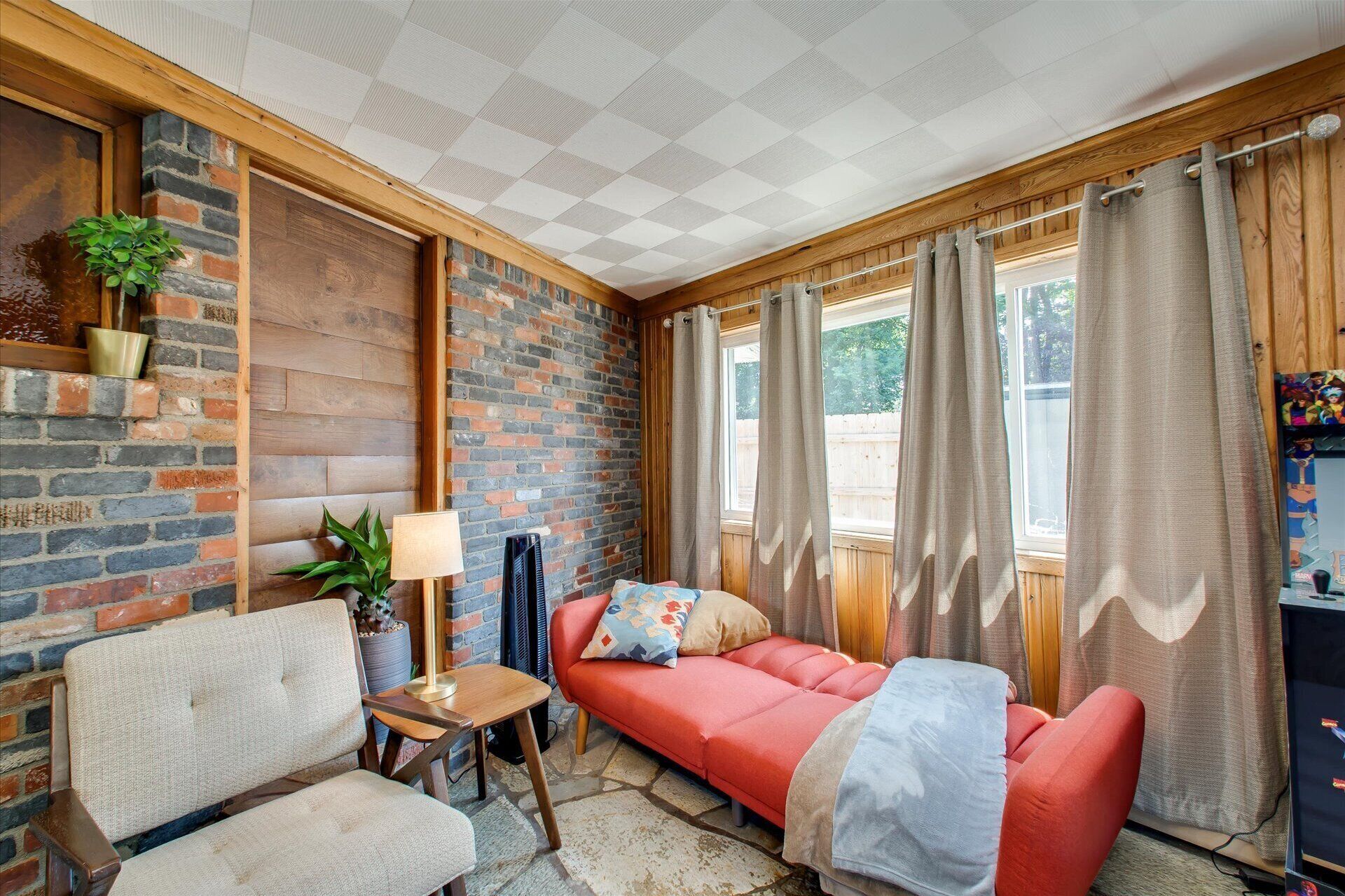 A cozy room with a brick wall, a wooden ceiling, a red sofa, a beige armchair, and a small table with a lamp.