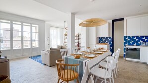 A spacious living room with a large window, a dining area with a striped tablecloth, and a kitchen with geometric backsplash.