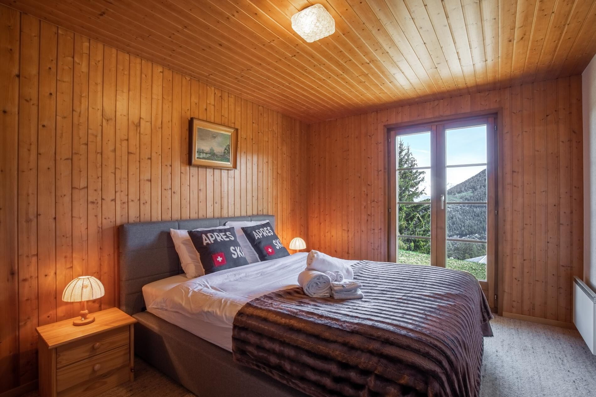 Een slaapkamer met houten lambrisering, een bed, nachtkastje en een raam met uitzicht op een berg.