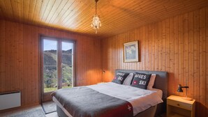 A wooden-paneled bedroom with a bed, a nightstand, a framed picture, and a view of a mountainous landscape through a large window. - Chalet Anne Sophie (Riddes)