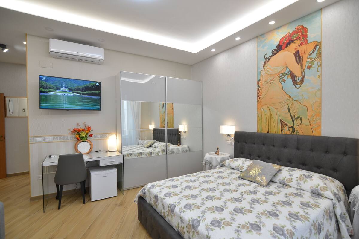 A modern bedroom with a bed, a desk, a chair, a small table, and a large mirror.