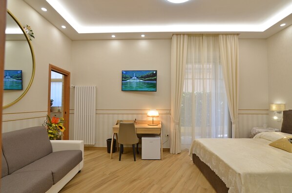 A hotel room with a bed, a desk with a lamp, a chair, a sofa, a small table with flowers, a mirror, and a picture on the wall. - Caserta Deluxe Bis (Caserta)