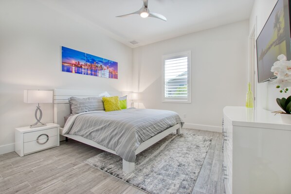A modern bedroom with a bed, nightstands, a ceiling fan, a window with blinds, and a painting on the wall. - Intervillas Florida - Villa Splendid (Cape Coral)