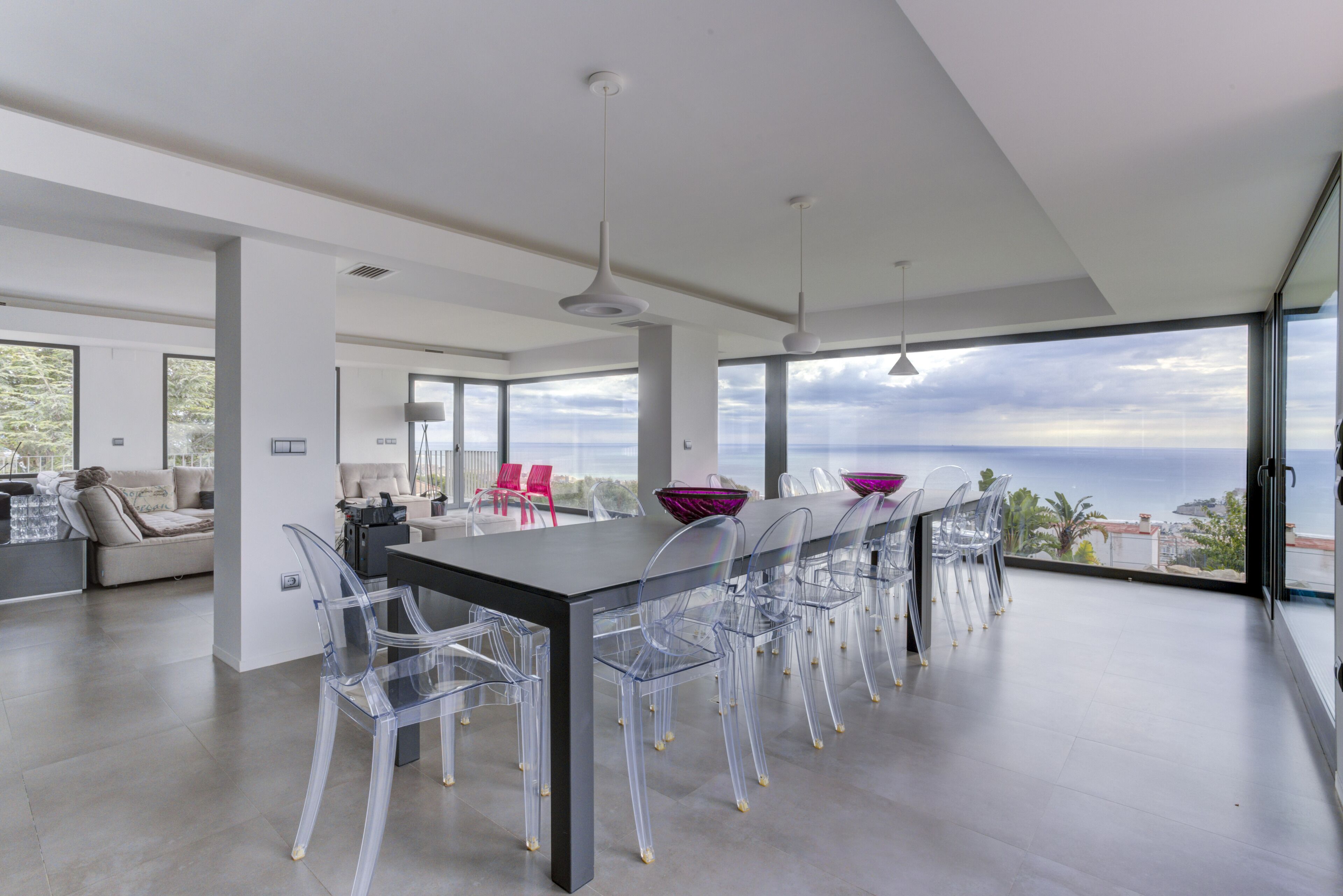 A modern dining area with a long table and clear chairs, large windows offering a view of the sea, and a minimalist interior design.