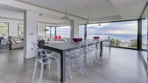 A modern dining area with a long table and clear chairs, large windows offering a view of the sea, and a minimalist interior design. - Villa Atalaya (Peñíscola)
