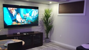 Ein modernes Wohnzimmer mit einem an der Wand befestigten Flachbildfernseher, einem Essbereich mit Tisch und Stühlen, einer großen Topfpflanze und einem grauen Teppich.