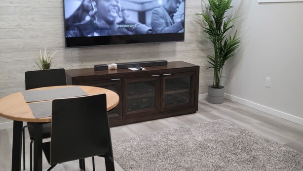 Ein modernes Wohnzimmer mit einem flachbildfernseher an einer hellen Wand, einer dunklen Holzunterhaltungsecke darunter, einem runden Holztisch mit schwarzen Stühlen und einem grauen Teppich.