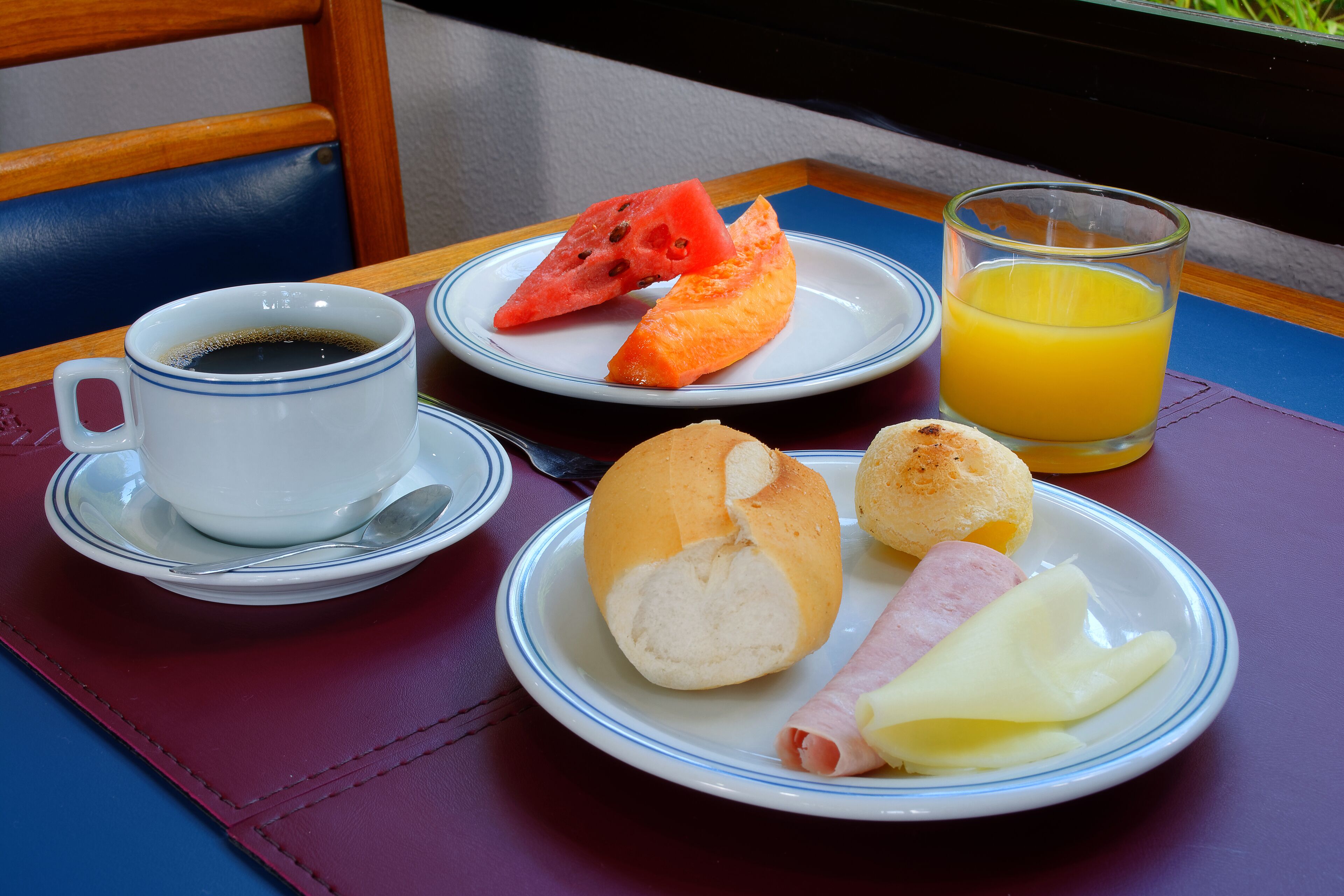 A breakfast table with a cup of coffee, a plate with bread, ham, cheese, and a slice of watermelon, and a glass of orange juice.