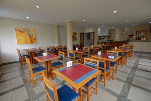 Un espace repas avec plusieurs tables et chaises, une cuisine à l’arrière-plan et un tableau accroché au mur.