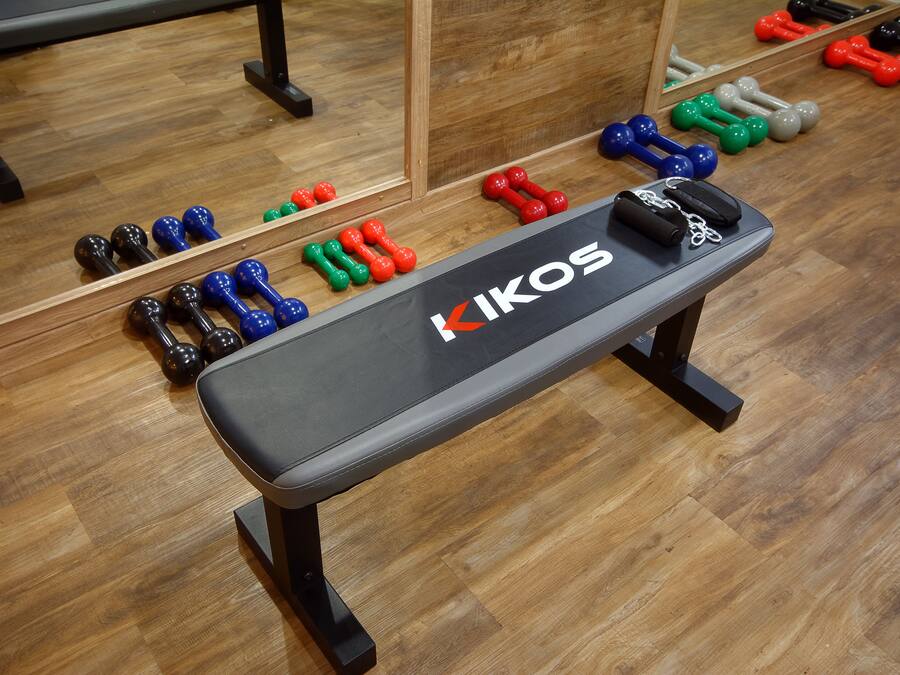 Un gimnasio con una banca con el rótulo "KIKOS" y varias mancuernas de colores dispuestas en el suelo.