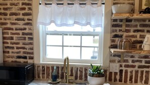 A kitchen with a brick wall, a window with a white valance, a sink, a microwave, and a potted plant. - Modern, cozy, Studio Apartment (Ochlocknee)