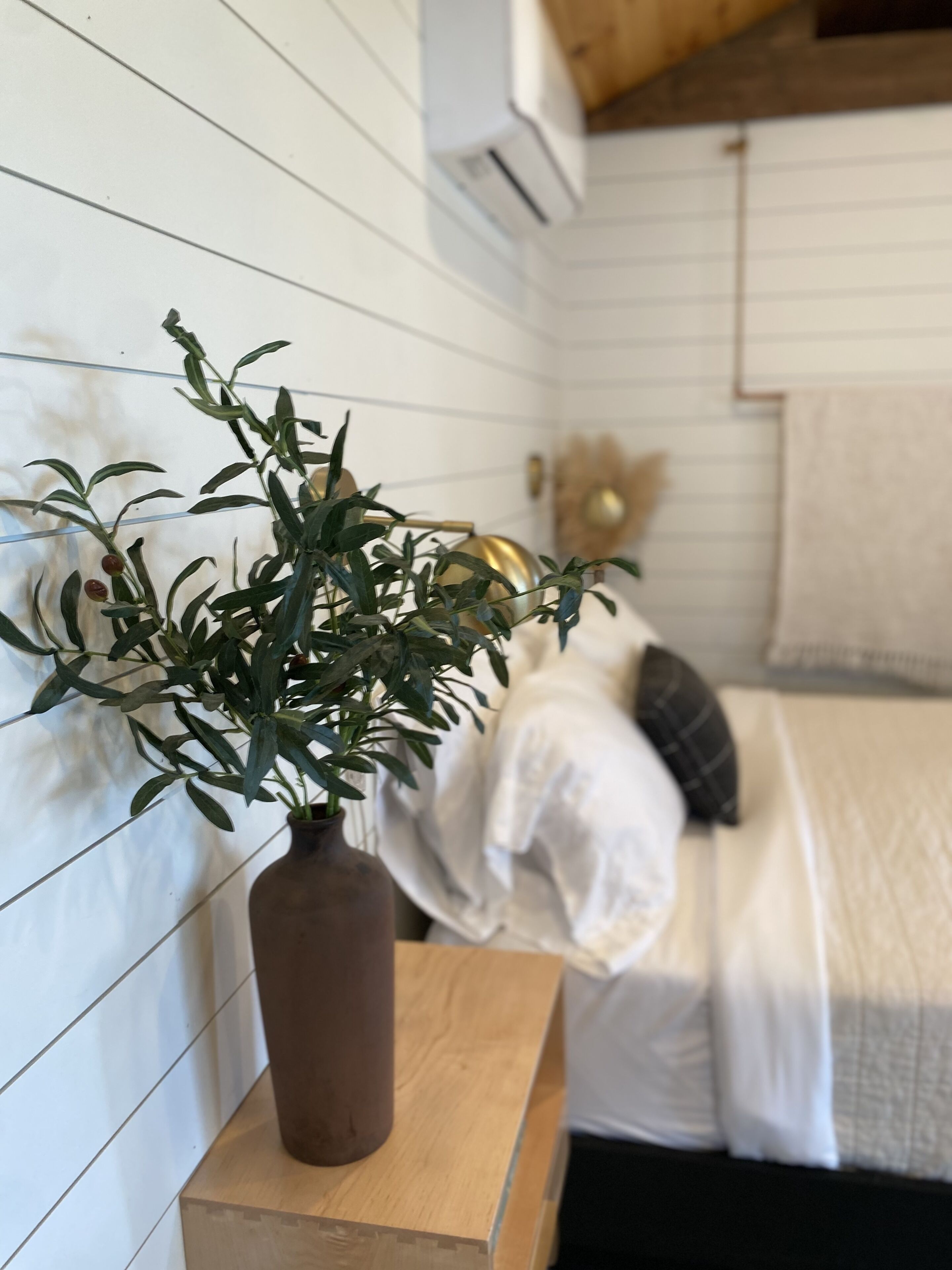 Una habitación con una mesita de noche de madera que sostiene un florero con una planta, una cama con ropa blanca y un adorno colgado en la pared.