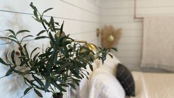 Una habitación con una mesita de noche de madera que sostiene un florero con una planta, una cama con ropa blanca y un adorno colgado en la pared.