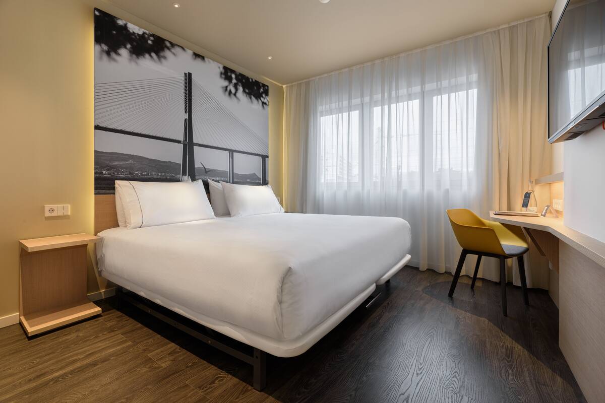 A modern hotel room with a large bed, a desk with a chair, and a large window with sheer curtains.