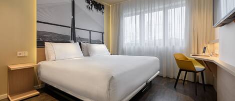 A modern hotel room with a large bed, a desk with a chair, and a large window with sheer curtains.