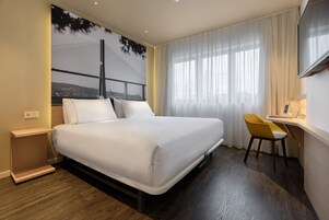 Une chambre d’hôtel moderne dotée d’un grand lit, d’un bureau avec une chaise et d’une grande fenêtre avec des rideaux légers.