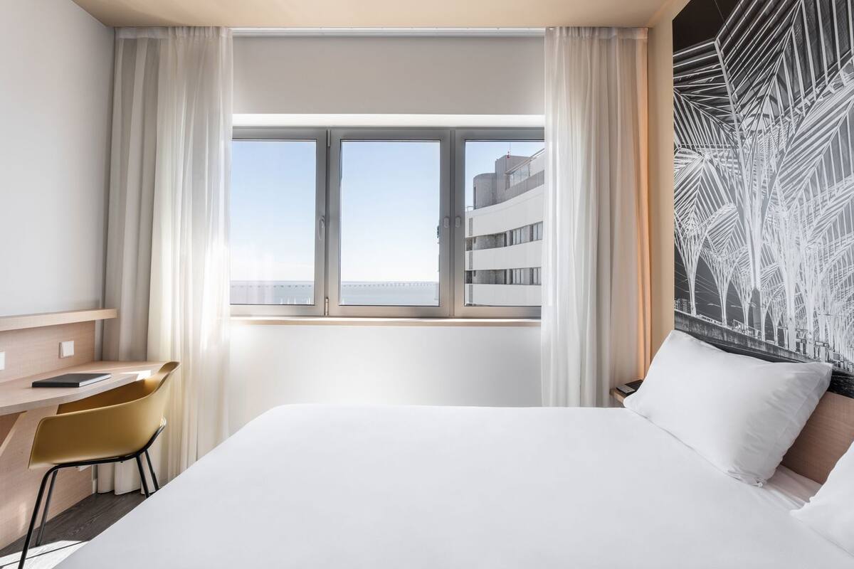 A hotel room with a bed, a desk, a chair, and a window with a view of buildings.