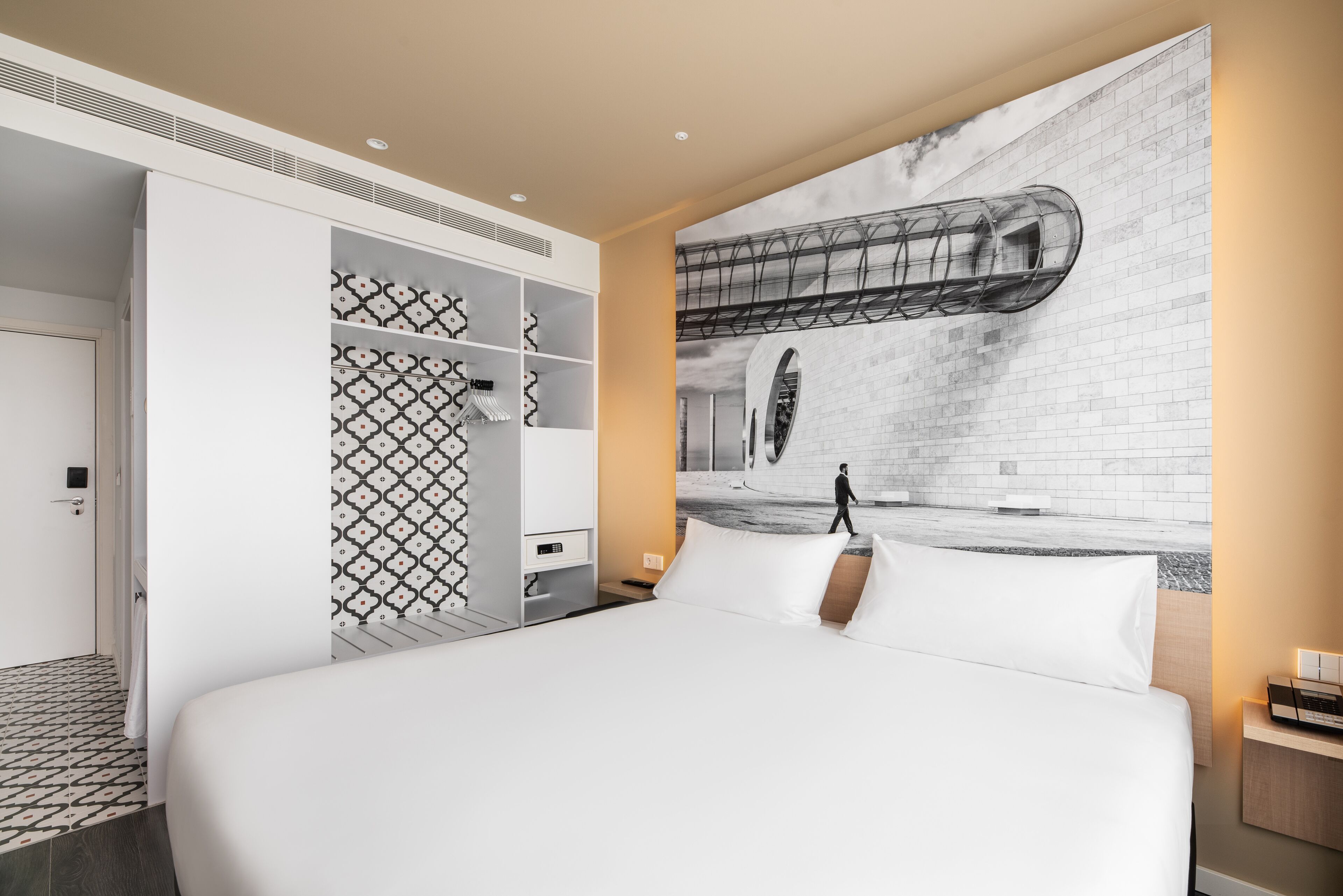 A modern hotel room with a large bed, a wardrobe with patterned doors, and a mural of a building on the wall.