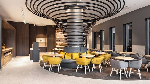 A modern interior with a central spiral column, seating areas with yellow and grey chairs, and a bar counter.