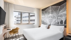 A modern hotel room with a large bed, a desk with a chair, a large window, and a unique wall art piece.