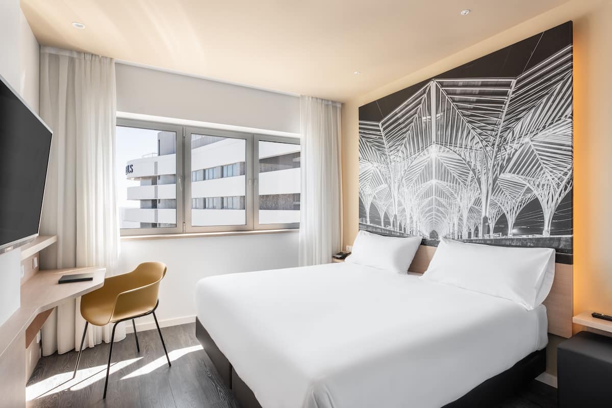 A modern hotel room with a large bed, a desk with a chair, a large window, and a unique wall art piece.