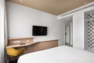 Moderni hotellihuone, jossa on sänky, työpöytä tuolilla, taulutelevisio ja vaatekaappi kuvioiduilla ovilla.