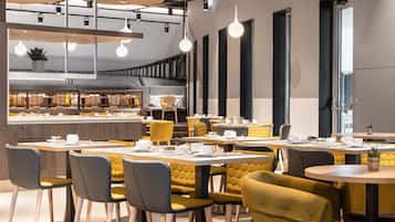 Un área de comedor moderna con mesas y sillas redondas, una cocina al fondo e iluminación colgante.