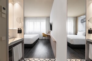 A hotel room with two beds, a desk, a chair, a TV, and a bathroom with a mirror and sink.