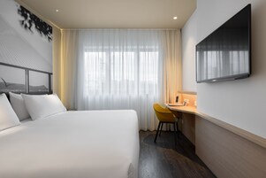 A hotel room with a bed, a desk, a chair, a TV, and a window with curtains.