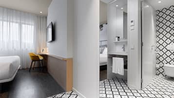 Quarto de hotel moderno com cama, escrivaninha, cadeira e banheiro com piso estampado, além de área de chuveiro.