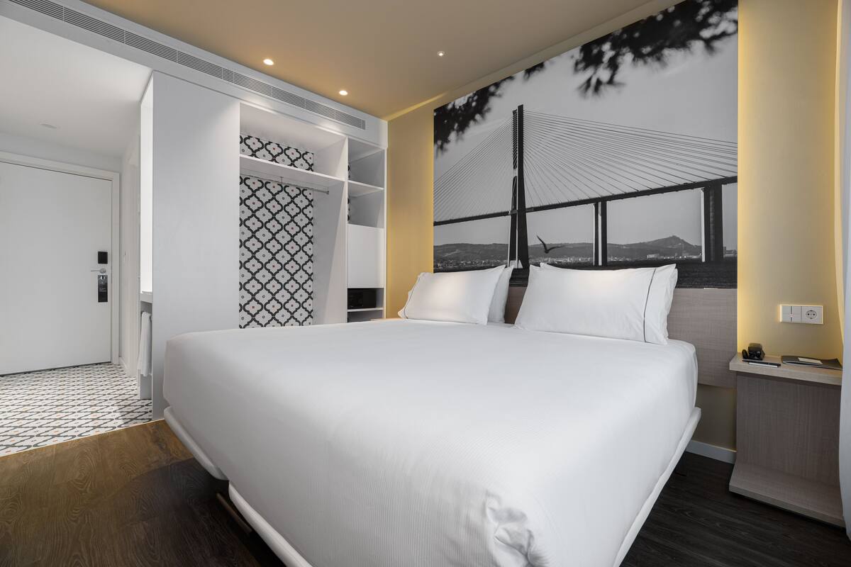 A modern hotel room with a large bed, a wardrobe, a desk, and a mural of a bridge.