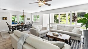 A spacious living room with a high ceiling fan, large windows offering a view of the outdoors, and a dining area with a table and chairs. -  Above Asheville, Art House  (Fairview)