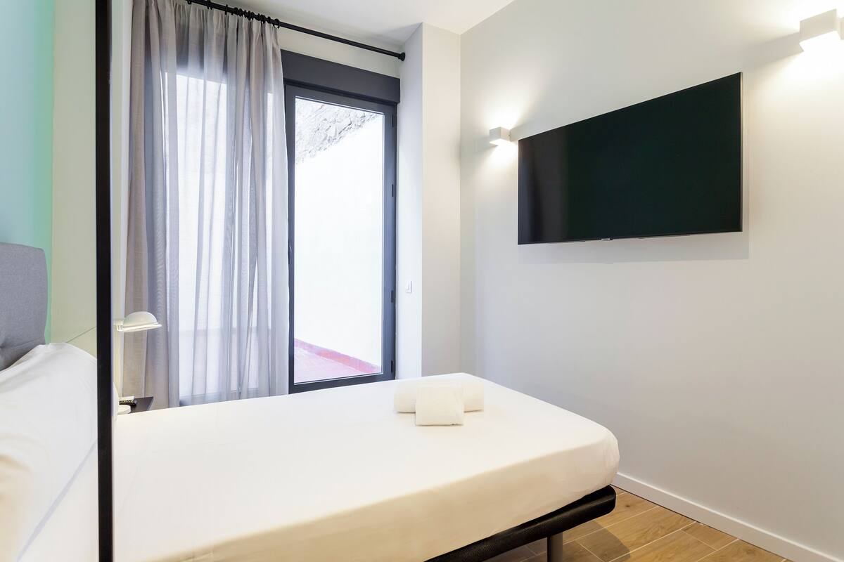 Quarto de hotel com uma cama, uma televisão presa à parede e uma janela com cortinas.