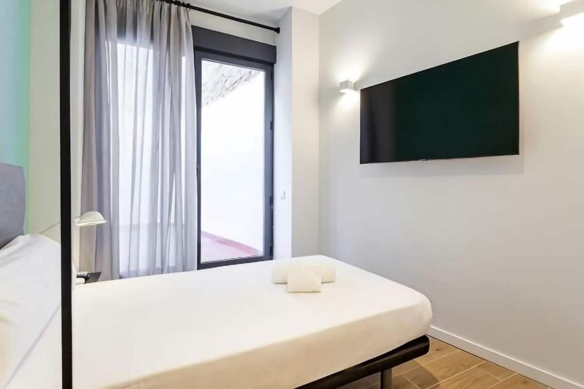 Quarto com cama, janela com cortinas, TV montada e porta visível.