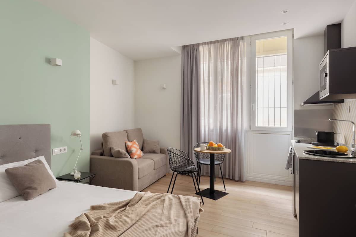 Quarto de hotel moderno com cama, sofá, mesa de jantar com cadeiras, cozinha e uma janela grande com cortinas.