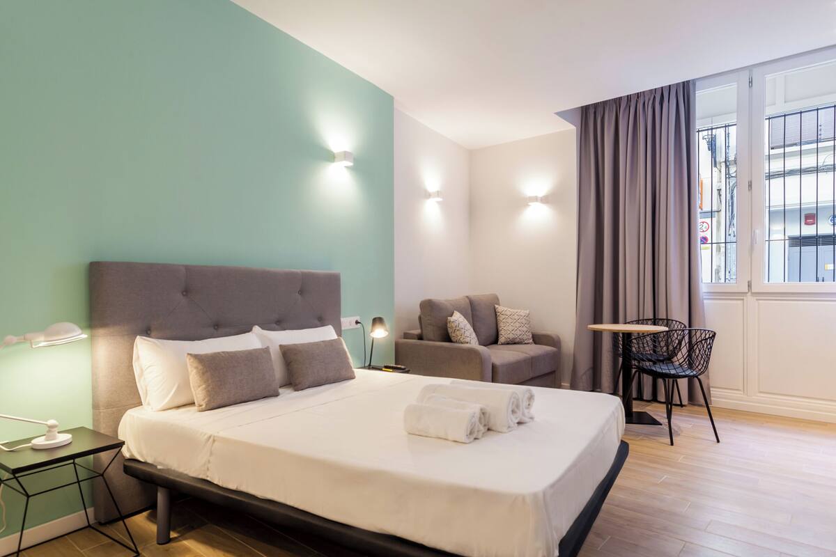Quarto de hotel moderno com uma cama grande, área de estar com sofá e uma mesinha, e vista pela janela.
