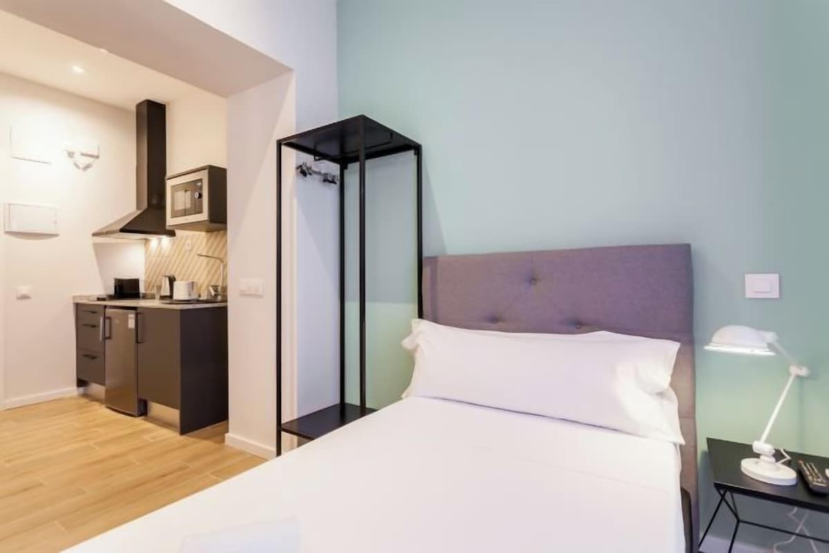 Una habitación de hotel moderna con cama, mesita de noche, lámpara y una zona de kitchenette.