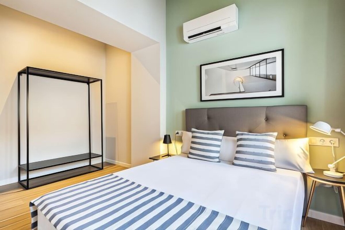 A hotel room with a bed, bedside lamps, a nightstand, a framed picture on the wall, and an air conditioning unit.